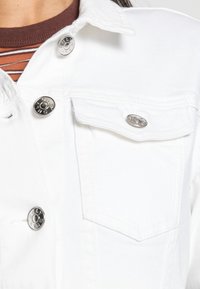 Vit denimjacka med silverfärgade tryckknappar och bröstficka. Tyget har en slät yta med en ren, minimal design.