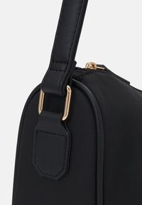 Schwarze Nylon-Tasche mit strukturierter Form, ausgestattet mit einem glatten Ledertragegriff und goldenen Hardware-Details. Der Reißverschluss ist oben sichtbar.