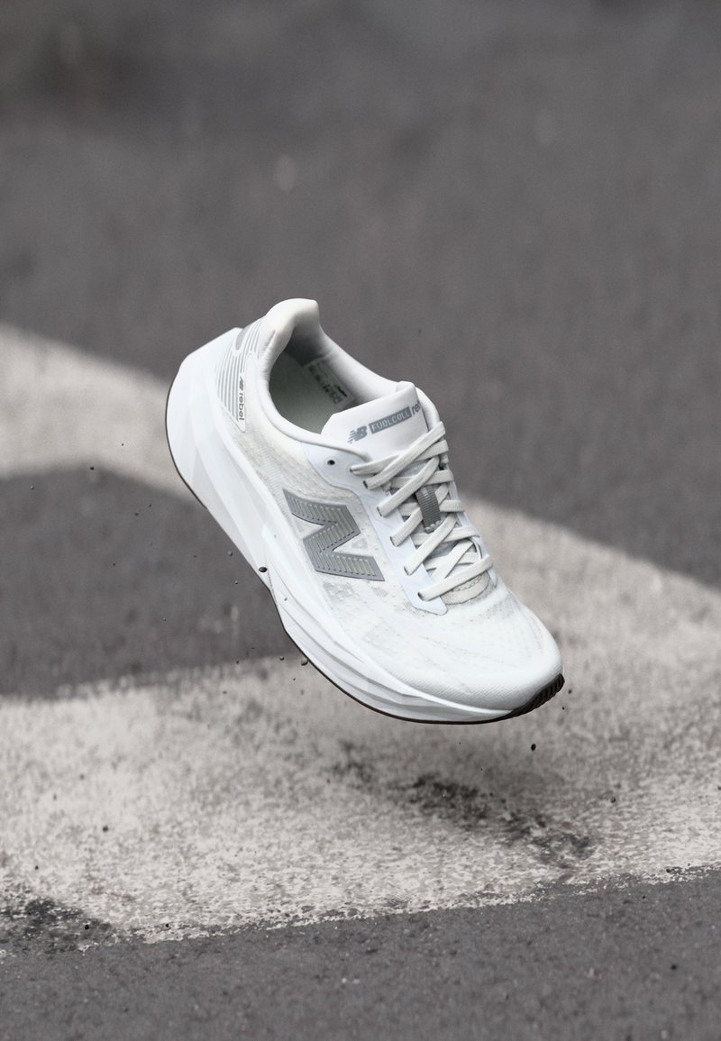 Chaussure de course blanche avec une tige en mesh texturé, accents gris et semelle rembourrée, suspendue dans les airs au-dessus d'un marquage au sol.
