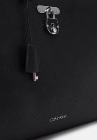 Černá kožená kabelka s texturou, stříbrným zámkem, detailem klíče a vytištěným logem. Disponuje hladkým zipem a strukturovaným tvarem.