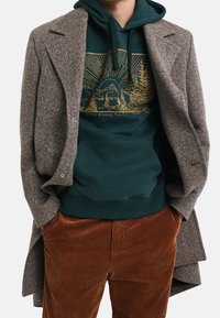 Abrigo de mezcla de lana gris superpuesto sobre una sudadera con capucha verde que presenta un gráfico dorado de montañas y árboles, combinado con pantalones de pana marrones.