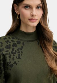 Donna con lunghi capelli castani che indossa orecchini a cerchio d'oro e un maglione verde con stampa leopardata e collo alto.