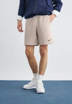 Άτομο που φορά μπεζ σορτς Nike με το λογότυπο στο αριστερό πόδι, λευκές κάλτσες και λευκά αθλητικά παπούτσια, στέκεται σε μπλε χαλί με το ένα χέρι στην τσέπη.