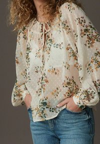 Femme portant un blouse florale blanche transparente avec de longues manches bouffantes et un jean bleu, les mains dans les poches, les cheveux bouclés partiellement visibles.