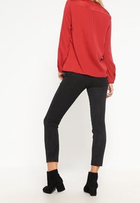 Blouse rouge à coupe décontractée, assortie à un pantalon noir à pois. Le modèle porte des bottines à talons noires. Texture du tissu lisse visible.