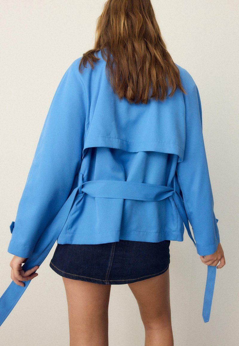 Femme aux longs cheveux bruns porte une veste bleu clair et une mini-jupe en denim foncé, tenant les lanières de la ceinture de la veste derrière son dos.