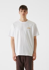 Bijela pamučna majica s malim bež logotipom na lijevoj strani prsnog koša, klasičnog okruglog izreza i ležernog kroja. Uparena s tamnosmešnim hlačama.