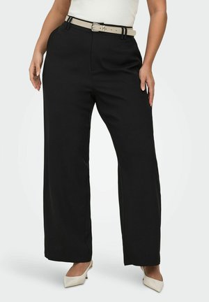 Femme portant un pantalon noir taille haute à jambes larges avec une ceinture beige et des escarpins beiges à bouts pointus, debout les mains détendues le long du corps.