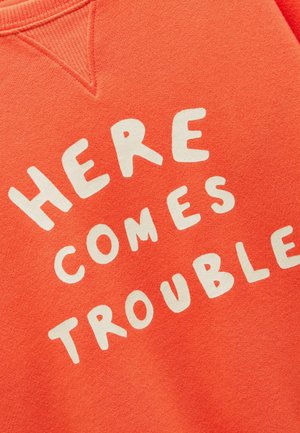 Nahaufnahme eines orangefarbenen Stoffes mit weißem Text "HERE COMES TROUBLE" in einer verspielten Schrift gedruckt.
