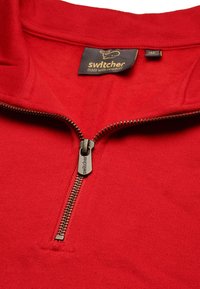 Roter Reißverschluss-Pullover aus weichem Stoff mit kurzem Kragen, goldfarbenem Reißverschluss und einem Etikett mit "Switcher"-Logo am Hals.