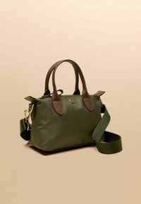 Zelená plátenná kabelka s hnedými koženými doplnkami, dvojitými rúčkami, odnímateľným remienkom, okrúhlym zlatým logom a horným zatváraním na zips.