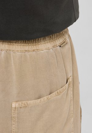 Béžové kalhoty s elastickým pasem a viditelným zadním kapsou, doplněné tmavým vrškem částečně viditelným v oblasti pasu.