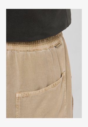 Beige broek met elastische tailleband en zichtbare achterzak, gecombineerd met een donkergekleurde top die gedeeltelijk zichtbaar is bij de taille.