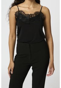 Svart camisole med intrikata svarta spetsdetaljer längs halsringningen, tunna justerbara band, i kombination med svarta skräddarsydda byxor.