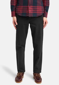 Pantaloni neri in cotone con vestibilità dritta, abbinati a scarpe maroon con lacci marroni. La camicia presenta un motivo a quadri rosso e blu navy.