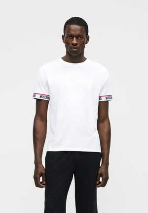 Mann trägt weißes T-Shirt mit kurzen Ärmeln und Moschino-Logo-Bändern an den Ärmeln sowie schwarze Hose, steht vor weißem Hintergrund.