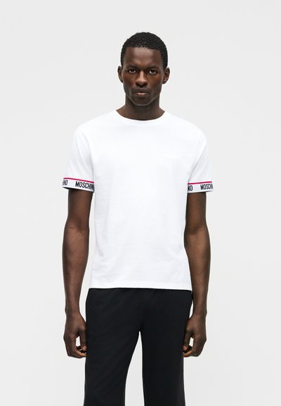Homme portant un t-shirt blanc à manches courtes avec des bandes logo Moschino sur les manches et un pantalon noir, debout devant un fond blanc uni.