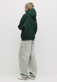 Felpa verde con un ampio cappuccio, abbinata a jeans oversize di colore grigio chiaro. Sneakers bianche completano l'outfit casual. Design minimalista e texture morbida.
