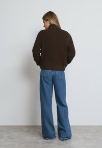 Pull à col montant côtelé marron associé à un jean en denim bleu loose, montrant l'arrière contre un fond clair.