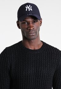 Cappellino da baseball blu navy con logo bianco ricamato, indossato con un maglione nero a maglia testurizzata caratterizzato da un colletto a girocollo e dettagli a coste.