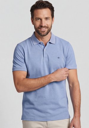 Man met kort donker haar en baard, gekleed in een lichtblauw poloshirt, die met zijn rechterhand de mouw rechttrekt tegen een neutrale achtergrond.