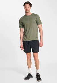 Oliwkowo-zielona sportowa koszulka z krótkimi rękawami i okrągłym dekoltem, w połączeniu z czarnymi szortami. Posiada subtelny geometryczny wzór. Czarne buty sportowe.