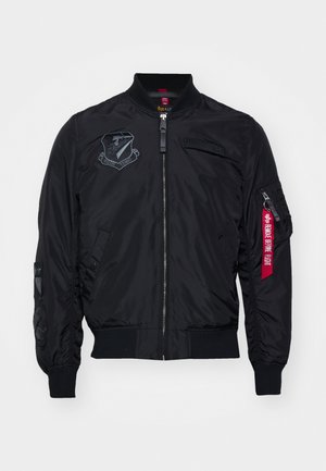 Veste bomber noire avec fermeture éclair à l'avant, poignets côtelés, écussons brodés et une étiquette en tissu rouge sur la manche. Matériau lisse et léger.