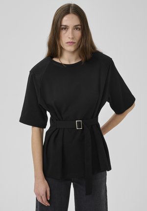 Fiatal nő hosszú barna hajjal, fekete öves rövid ujjú felsőt és sötét farmert visel, egy sima szürke háttér előtt áll.