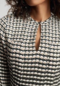 Schwarz-weiß gemusterte Bluse mit einem Schlüsselanschnitt-Ausschnitt, die geometrische Formen und Akzente in Orange aufweist. Glatte Stofftextur.