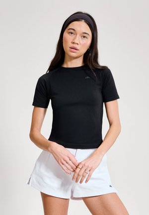 Mladá žena s dlouhými tmavými vlasy, oblečená v černém krátkém rukávu, bílých šortkách a černé čelence, stojící se sepjatýma rukama.