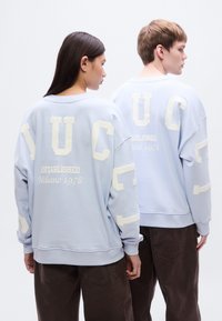 Φαρδιά φούτερ σε ανοιχτό μπλε χρώμα, με μείγμα βαμβακιού, διακοσμημένο με μεγάλες κρεμ γράμματα και την επιγραφή «Established Milano 1978» στην πίσω πλευρά.