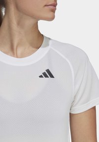 Balta sportinė marškinė su tekstūruota medžiaga, kairėje petyje esantis juodas "Adidas" logotipas. Apvalus iškirpimas ir trumpos rankovės.