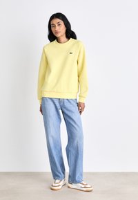 Mulher vestindo um sweatshirt amarelo pálido, calças jeans largas de tom azul claro e ténis brancos com detalhes em bege, apoiada contra um fundo simples.
