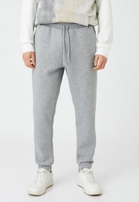 Grijze katoenen sweatpants met een elastische tailleband en trekkoord. Tapered pijpen met elastische boorden. Draagt met witte sneakers.