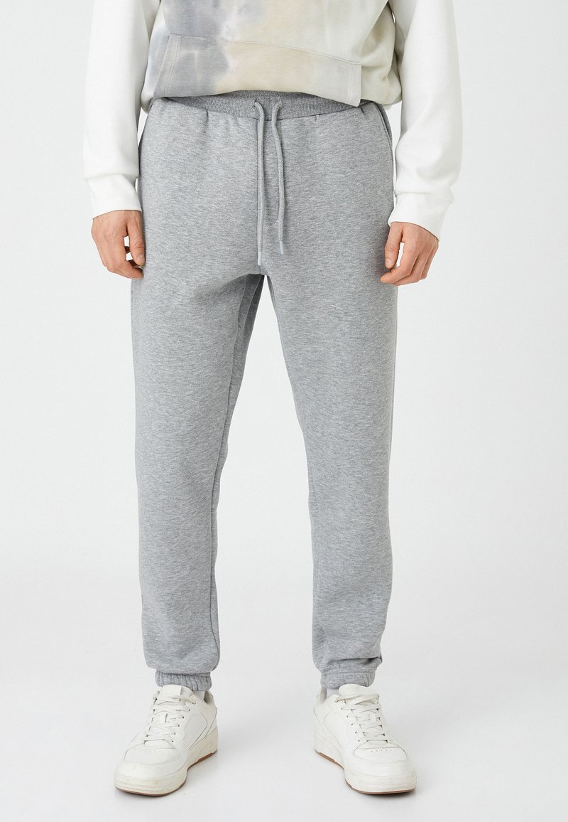 Grijze katoenen sweatpants met een elastische tailleband en trekkoord. Tapered pijpen met elastische boorden. Draagt met witte sneakers.