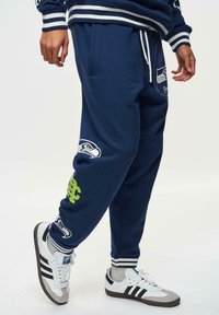 Marineblauwe sweatpants met gestreepte tailleband, voorzien van een Seahawks-logo en groene accenten. Gemaakt van zachte stof, ontworpen voor comfort en stijl.