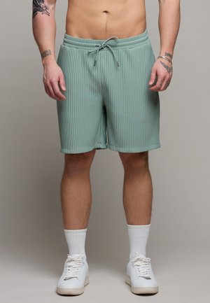 Mann trägt hellgrüne, gerippte Shorts mit Kordelzug, weiße Sportsocken und weiße Turnschuhe, steht vor einem schlichten Hintergrund.