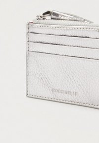 Zilveren leren kaarthouder met ritsvak, vier kaartsleuven en geperste "COCCINELLE" merknaam.