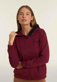 Sudadera con cremallera burdeos, capucha negra, mangas largas, bolsillos frontales y el texto "HOY ES TU DÍA" bordado en el pecho.