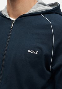 Close-up van een marineblauwe hoodie met ritssluiting, grijze voering en witte biezen, met een geborduurd "BOSS"-logo op de borst.