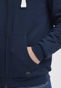 Granatowa bluza z kapturem na zamek, wykonana z miękkiego materiału, z białym sznurkiem, przednimi kieszeniami i ściągaczami przy mankietach, z metką umieszczoną w pobliżu dolnego brzegu.