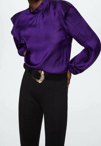 Lila Satinbluse mit gerafften Schultern, hohem Kragen und langen Ärmeln. Kombiniert mit schwarzen Hosen und einem mit Gold akzentuierten Gürtel.