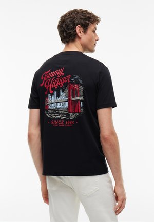 Man facing sideways wearing black Tommy Hilfiger t-shirt with red and white Brooklyn Bridge and city skyline design on back.