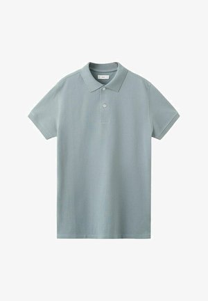 Junger Mann steht drinnen und trägt ein graues Poloshirt, dunkle Jeans, beige Schuhe und einen marineblauen Pullover, der über seine Schultern gelegt ist.