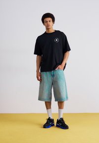 Černé oversized tričko s kulatým grafickým vzorem, světle modré vybledlé džíny, bílé ponožky a černé tenisky s modrými akcenty.
