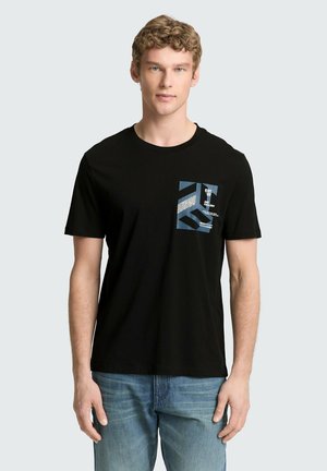 Jonge man met kort krullend haar die een zwart T-shirt met afbeelding en een blauwe spijkerbroek draagt, staat voor een effen lichte achtergrond.