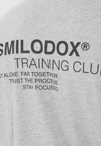 Heather grå t-shirt med sort tekst. Teksten inkluderer "SMILODOX® TRAINING CLUB" og motiverende sætninger. Blødt stof, korte ærmer.