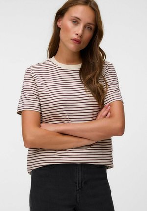 Jeune femme aux longs cheveux bruns porte une chemise blanche à manches courtes avec de fines rayures horizontales foncées et un pantalon noir, bras croisés.