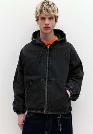 Mladá osoba v čiernej mikine na zips s kapucňou, s rukou v vrecku, stojaca pred jednoduchým bielym pozadím.