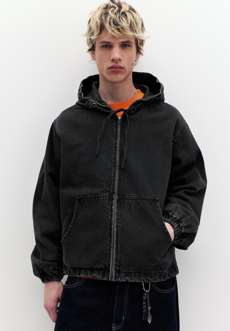 Jeune personne portant une veste à capuche noire avec fermeture éclair, main dans la poche, debout devant un fond blanc uni.